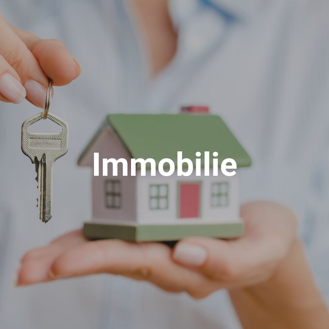 Eine Hand hält ein Modellhaus mit der Aufschrift "Immobilie" und eine Schlüsselkette.